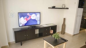 Ein modernes Wohnzimmer mit einem Flachbildfernseher an einem schwarzen Schrank, einem kleinen Holztisch mit einer Pflanze und einer Küchenzeile im Hintergrund.