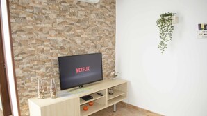 Olohuone, jossa on kiviseinä, televisio, jossa näkyy teksti »NETFLIX», puinen pöytä ja pieni hylly koriste-esineitä varten.