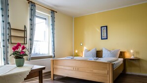 A bedroom with a wooden bed, a bedside table, a window with curtains, a radiator, a wall-mounted picture, and a bedside lamp. - Logia Hotel garni (Neuharlingersiel)