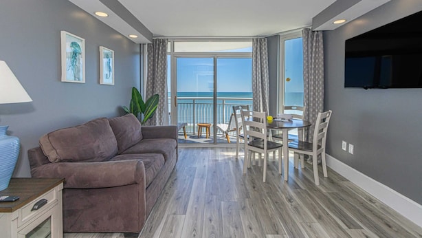 A modern living room with a sofa, a dining table with chairs, a TV, and a balcony with a view of the sea. - Atlantica 803 (Myrtle Beach)