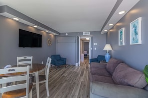 A modern living room with a flat-screen TV, a dining table with chairs, a sofa, and a hallway leading to another room.