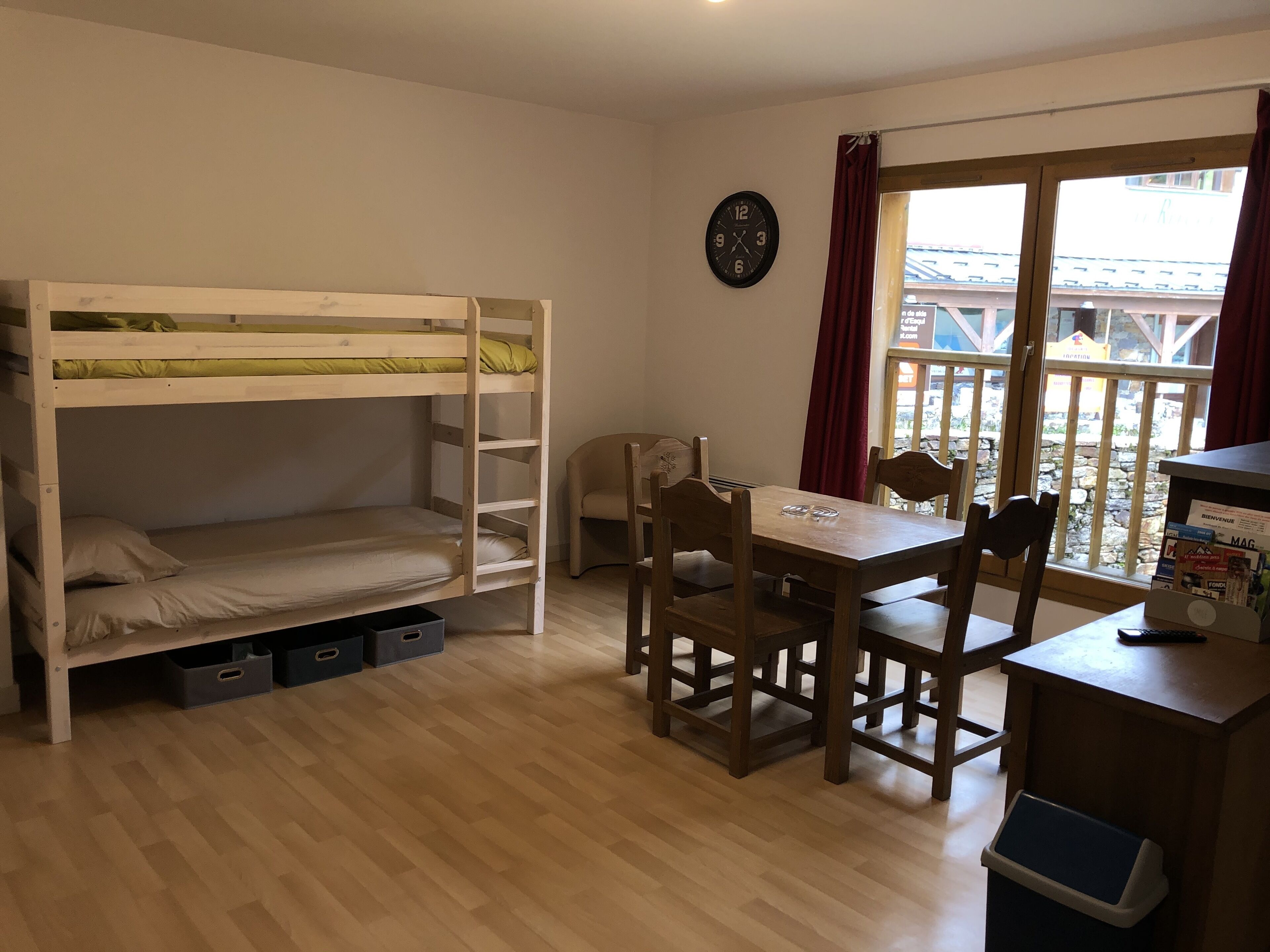Quarto com cama de solteiro, escrivaninha de madeira com cadeiras, relógio na parede e janela com cortinas.