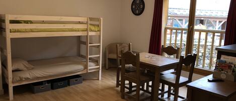 Quarto com cama de solteiro, escrivaninha de madeira com cadeiras, relógio na parede e janela com cortinas.