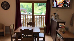A dining area with a wooden table and chairs, a television mounted on the wall, and a view of a wooden deck and greenery outside. - 4-person apartment, at the foot of the slopes, Ariège (Ustou)
