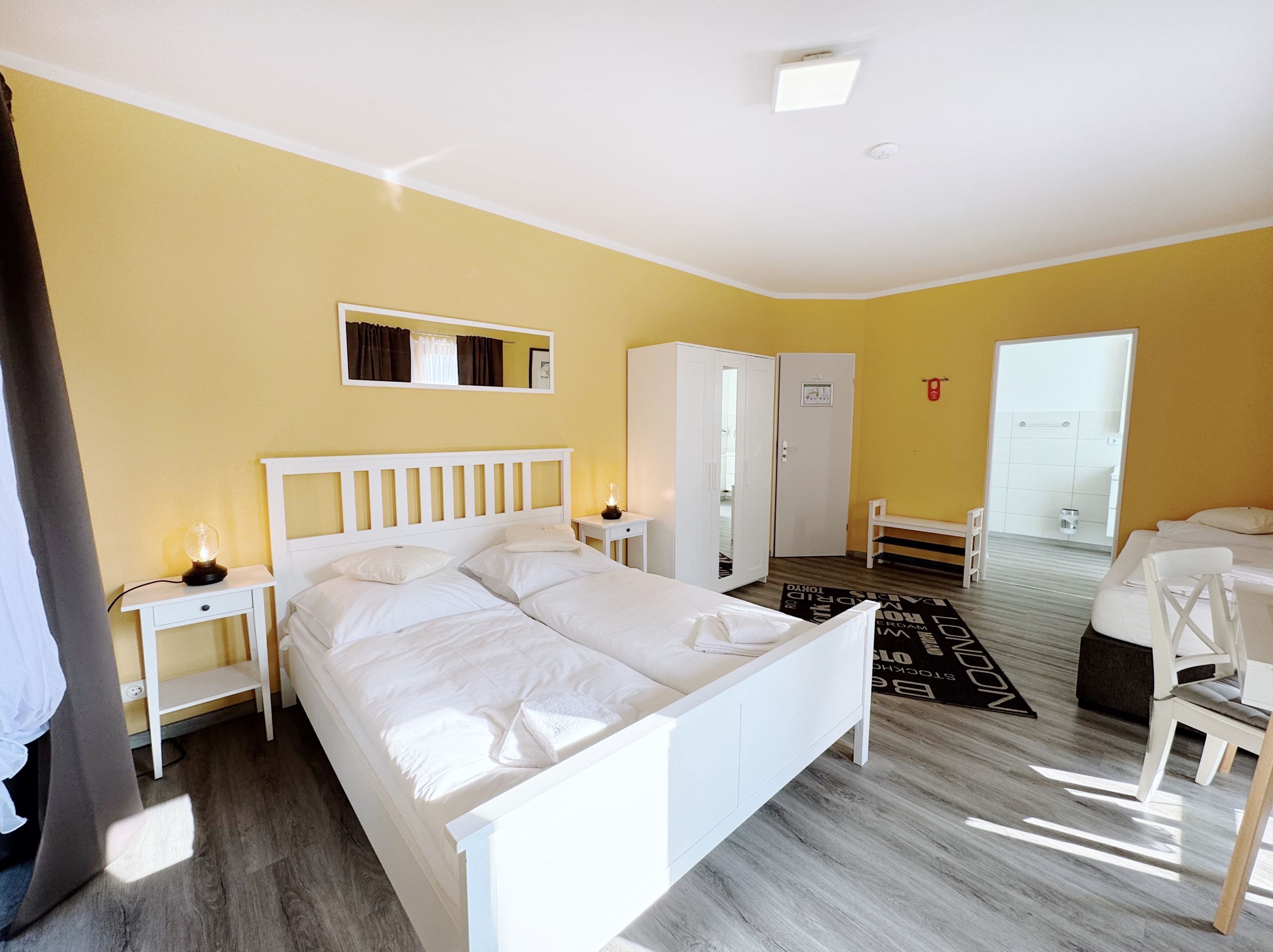 Habitación de hotel con cama doble, mesita de noche con lámpara, armario, banco y puerta que da a un baño.