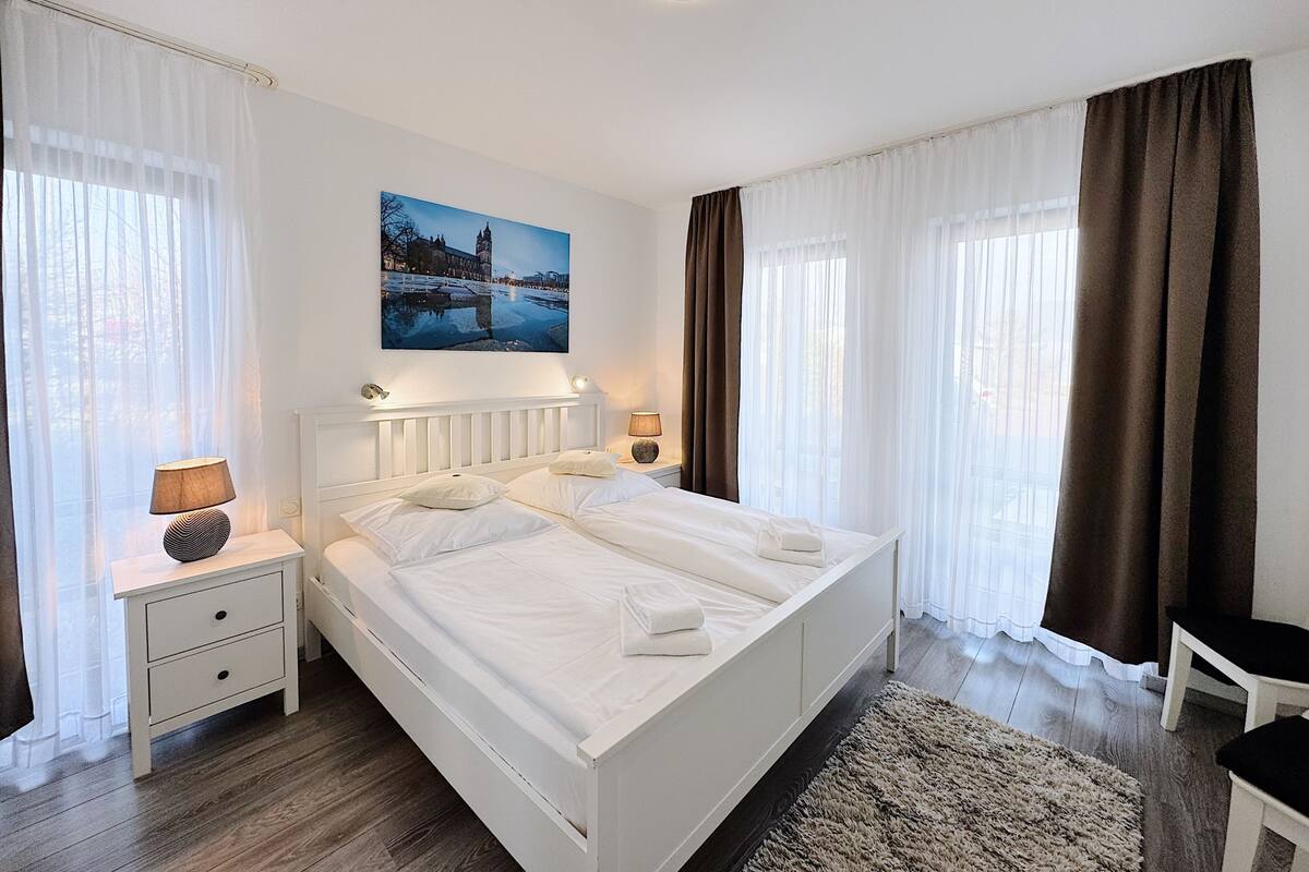 A spacious bedroom with a large bed, two bedside tables, a nightstand, and a view of a cityscape through sheer curtains.