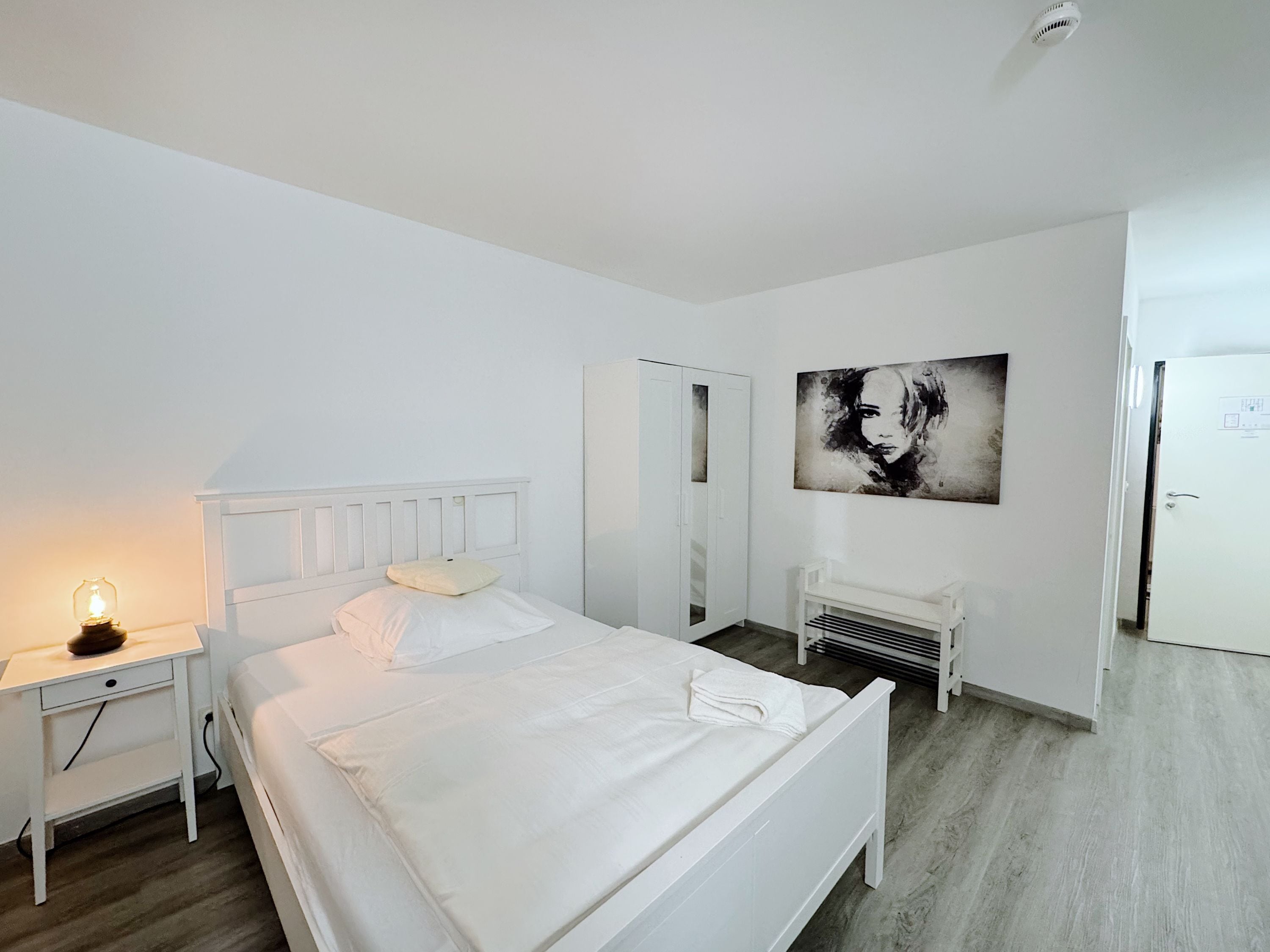 Un dormitorio blanco con cama, mesita de noche con lámpara, un banco, un armario y un cuadro en la pared.