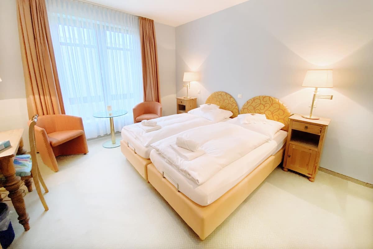 A hotel room with a double bed, two bedside tables with lamps, a chair, a small round table, and a desk with a chair.