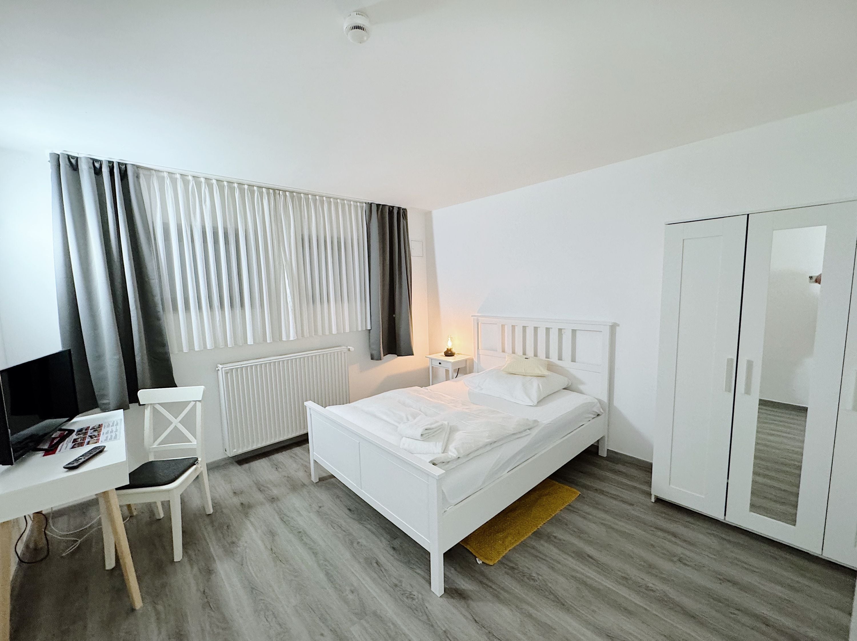Ein Schlafzimmer mit einem Bett, einem Schreibtisch mit Stuhl, einem Heizkörper, einem Kleiderschrank und einem Fenster mit Vorhängen.