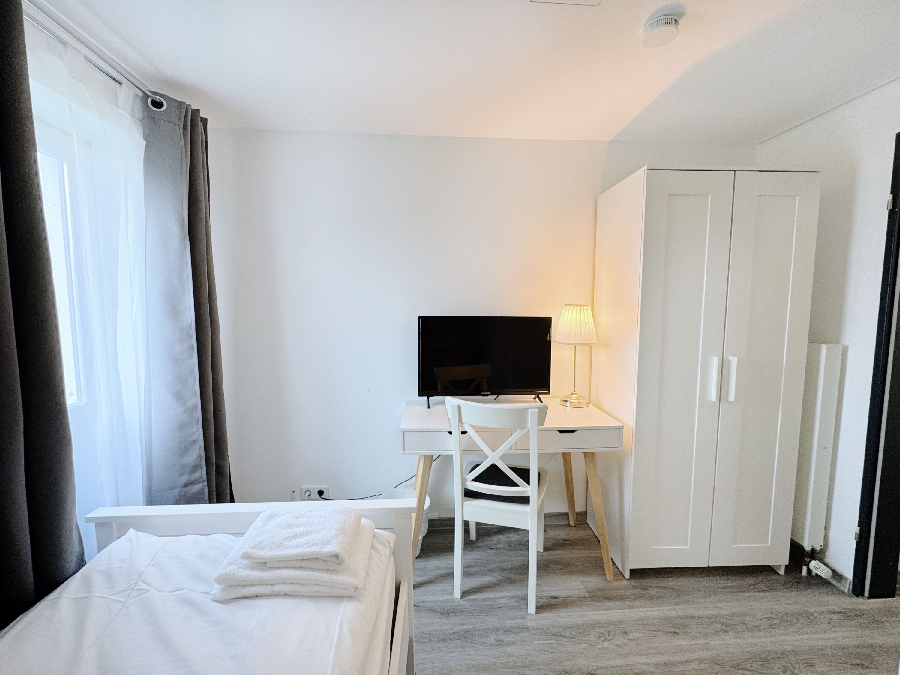 A hotel room with a bed, a desk with a TV, a chair, and a wardrobe.