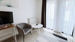 Un saló modern i minimalista amb televisió plana, un armari gris, una taula blanca petita i una finestra gran amb cortines primes.