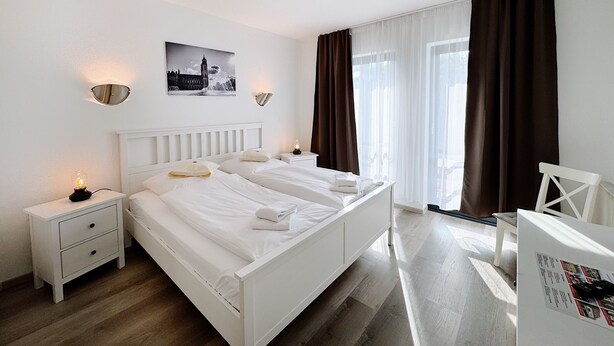 Habitació d'hotel amb llit blanc, taules de nit, escriptori i una cadira. Hi ha un quadre a la paret i una finestra amb cortines.