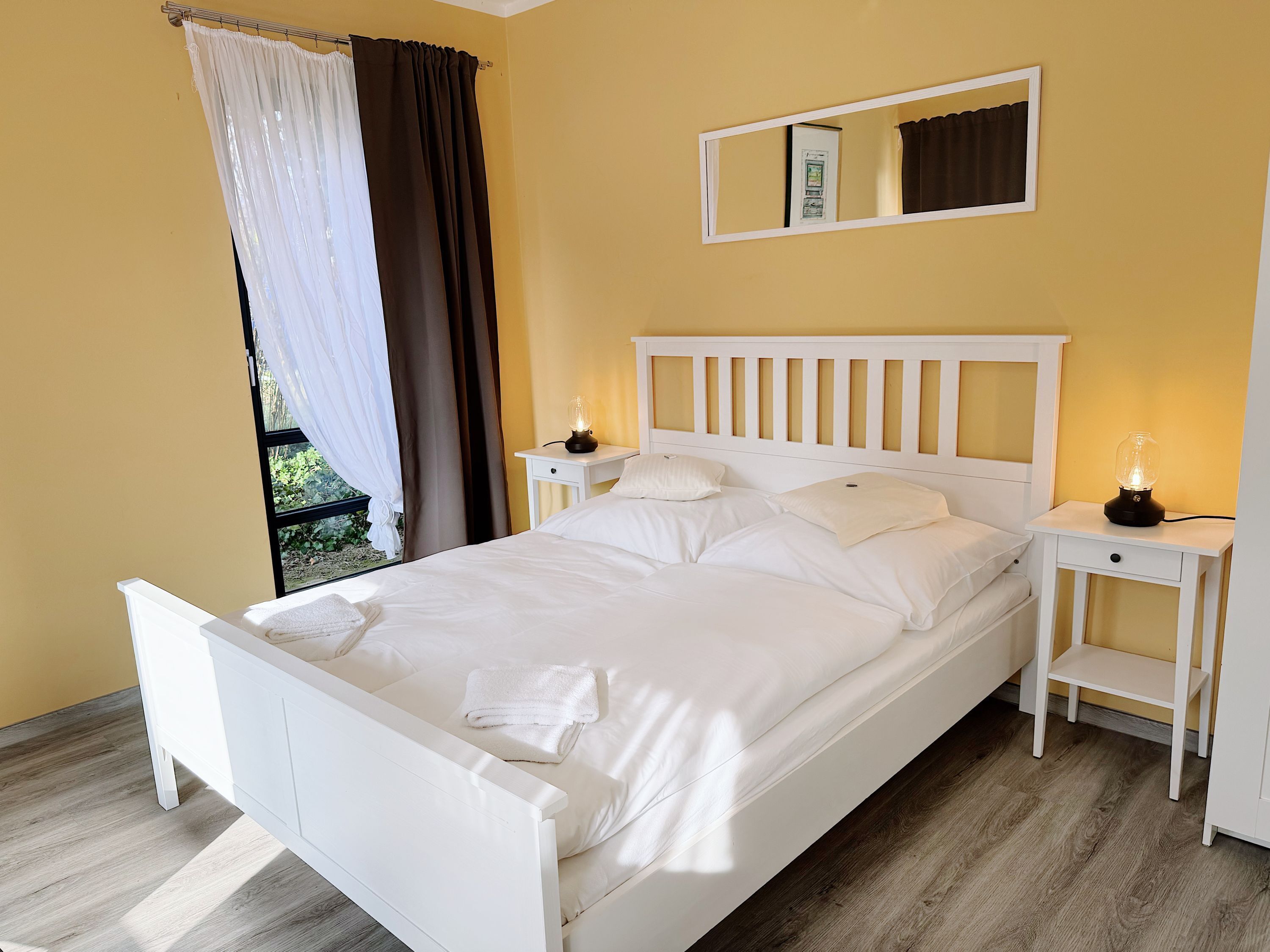 Una cama bien hecha con sábanas blancas, dos mesitas de noche con lámparas, una ventana con cortinas y un espejo en la pared.