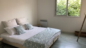 Cama bem arrumada com roupa de cama estampada, uma pequena mesa de cabeceira, um radiador e uma janela com vista para o verde.