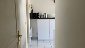 Um corredor estreito com piso ladrilhado que leva a uma área de cozinha com armários brancos e bancada preta.