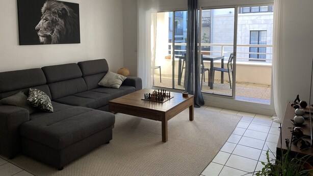 Uma sala de estar moderna com um sofá cinza escuro, uma mesa de centro de madeira e um jogo de xadrez. Há uma janela grande com vista para uma varanda externa.