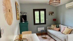 Ein Wohnzimmer mit Esstisch, einer Couch, einem Fernseher auf einem Standfuß, einem Fenster und einer Klimaanlage.
