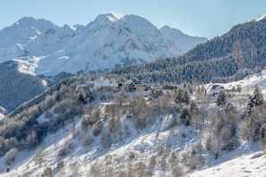 A snow-covered mountain village with chalet-style buildings, surrounded by forested slopes and snow-capped peaks.