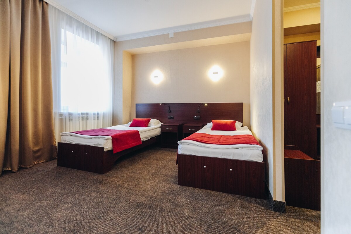 A hotel room with two beds, a large window with curtains, a wooden headboard, and a door to a closet.
