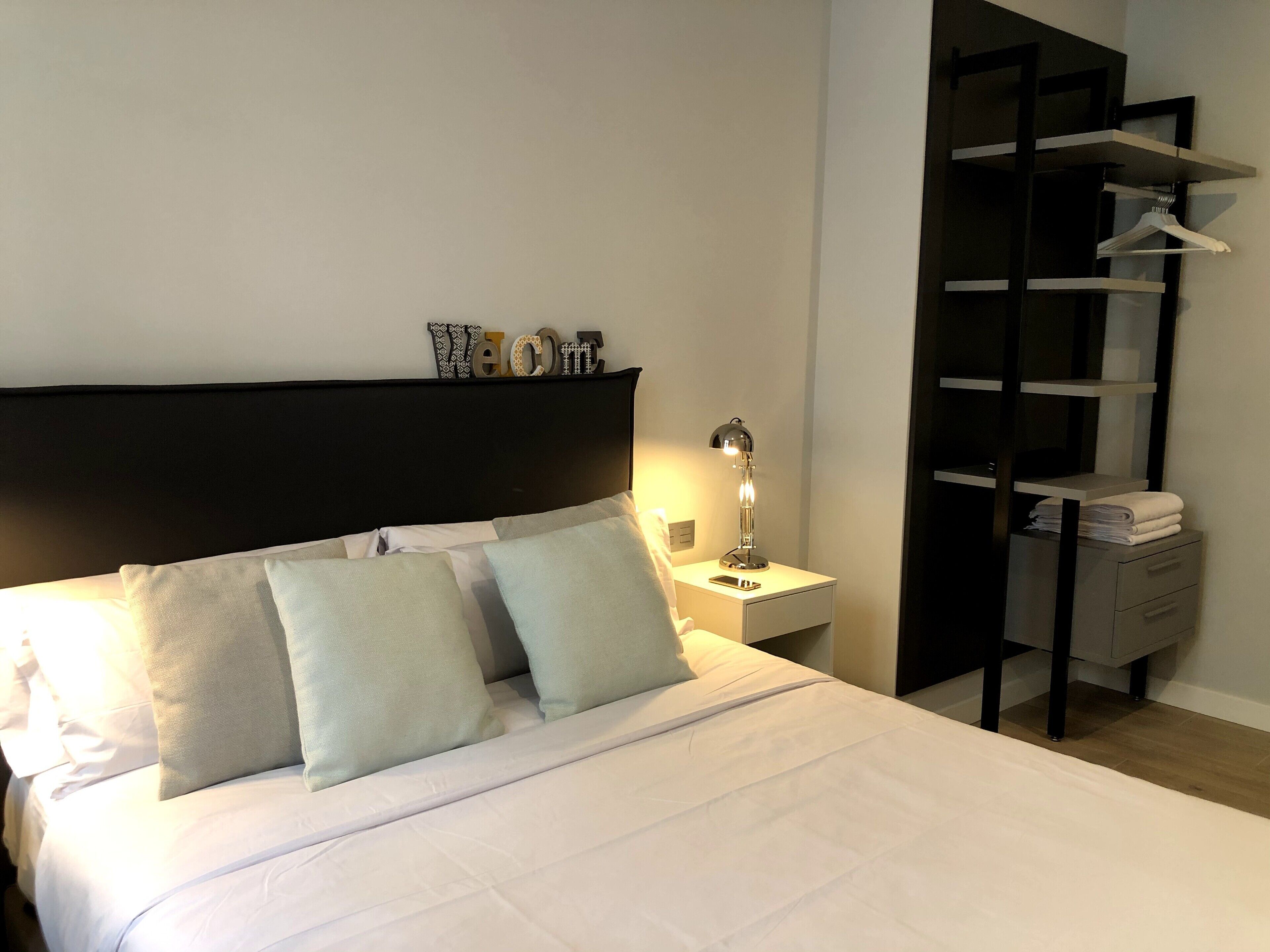 Una cama bien hecha con almohadas, una lámpara de noche y una mesita con una toalla doblada y un teléfono.