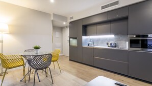 Uma cozinha moderna com armários cinza, micro-ondas de aço inoxidável e uma cafeteira. Há uma mesa de jantar com duas cadeiras, uma amarela e outra cinza. Um controle remoto está sobre a mesa.