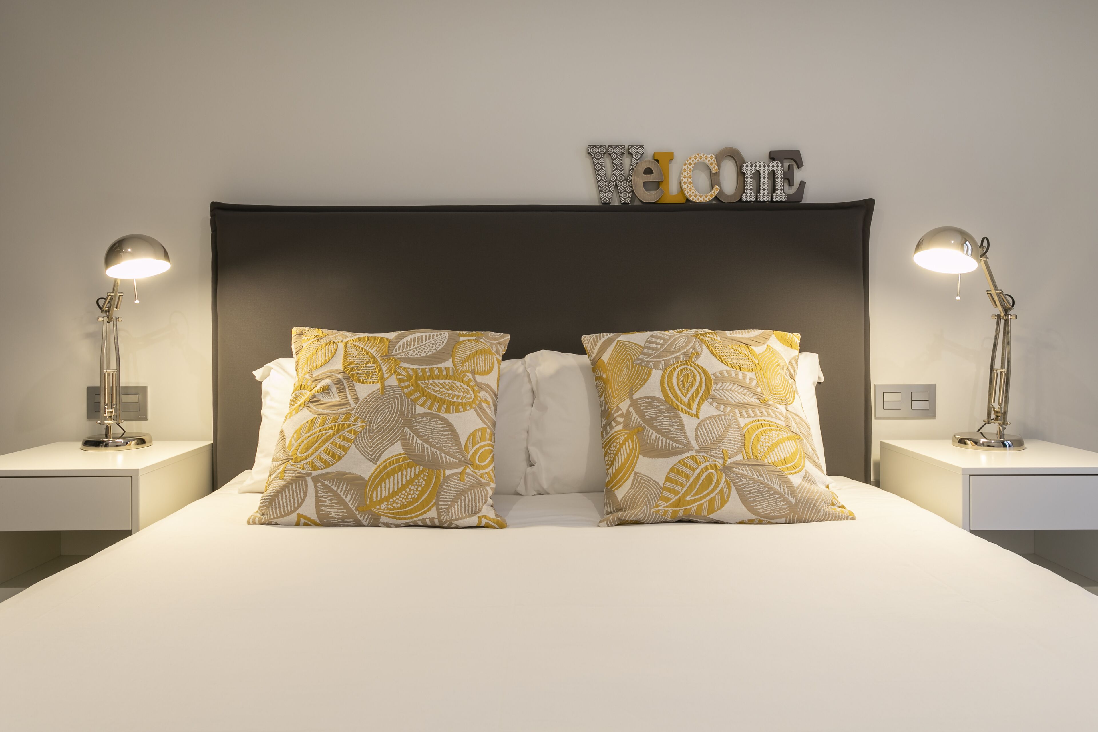 Una cama bien hecha con almohadas estampadas, cabecera oscura y dos lámparas de noche.