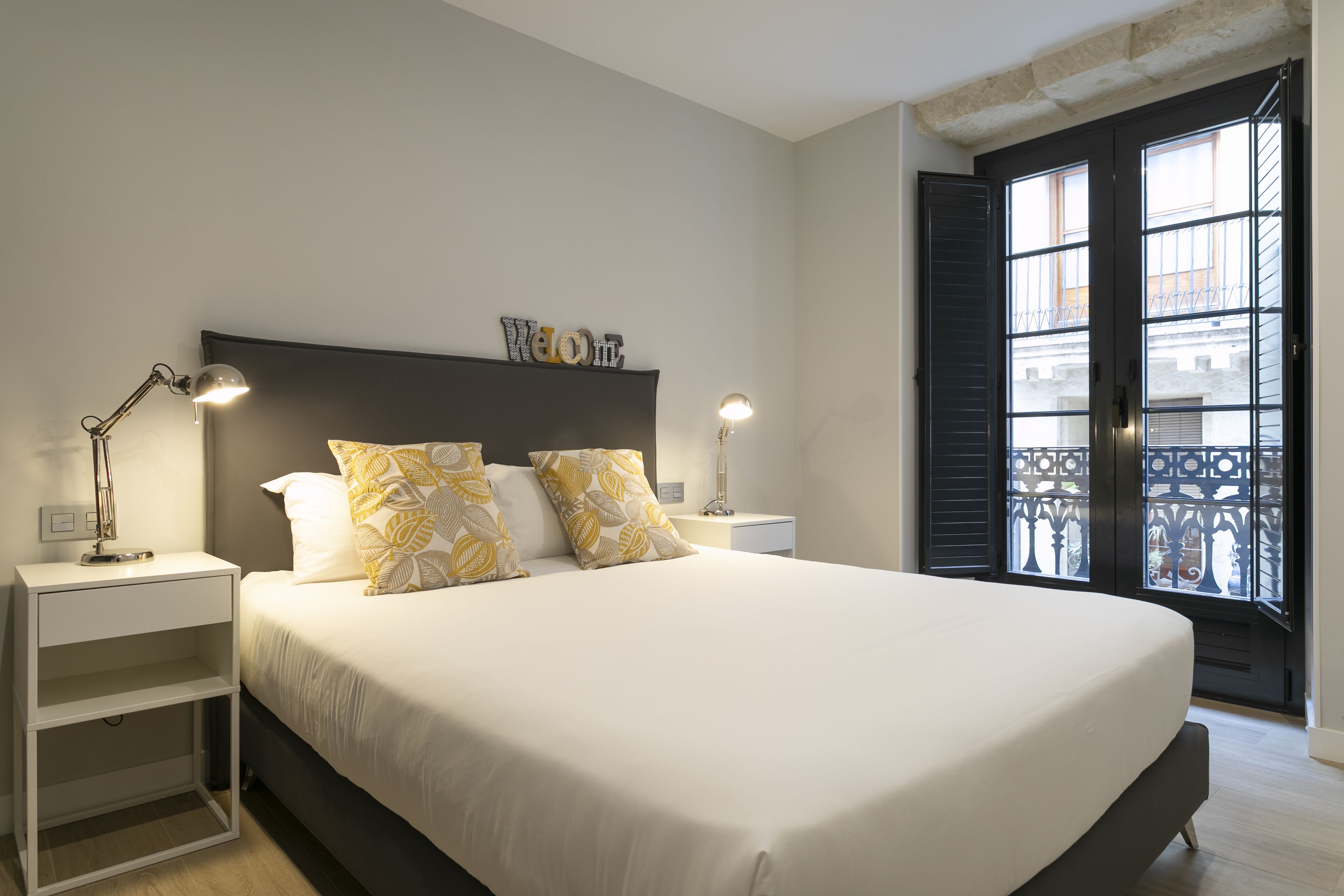 A neatly made bed with white bedding and two decorative pillows, a bedside table with a lamp, and a "Welcome" sign above the bed.