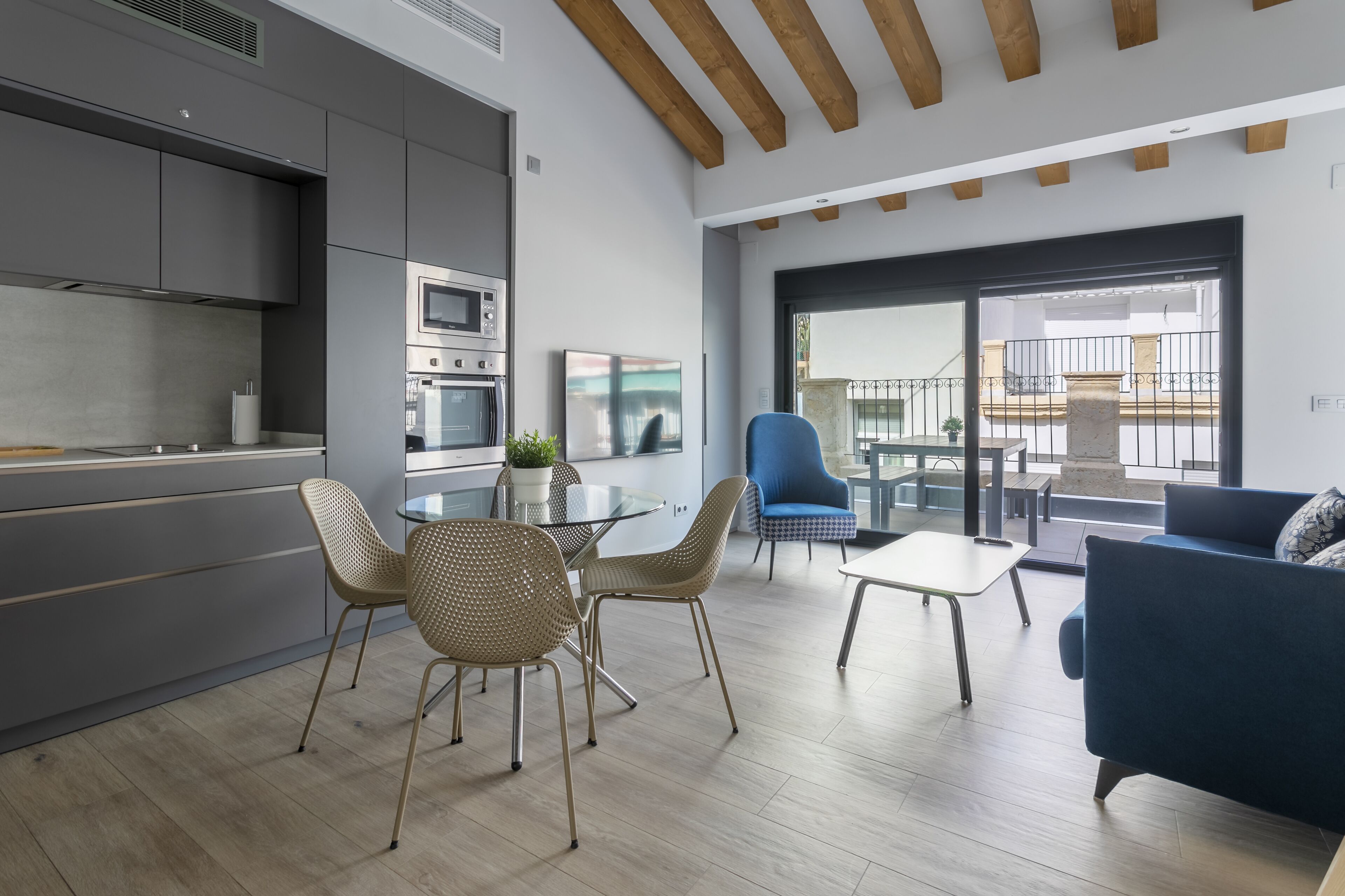 Eine moderne Küche mit integrierten Geräten, ein Essbereich mit Glastisch und Korbstühlen sowie ein Wohnbereich mit blauem Sofa und kleinem Tisch.