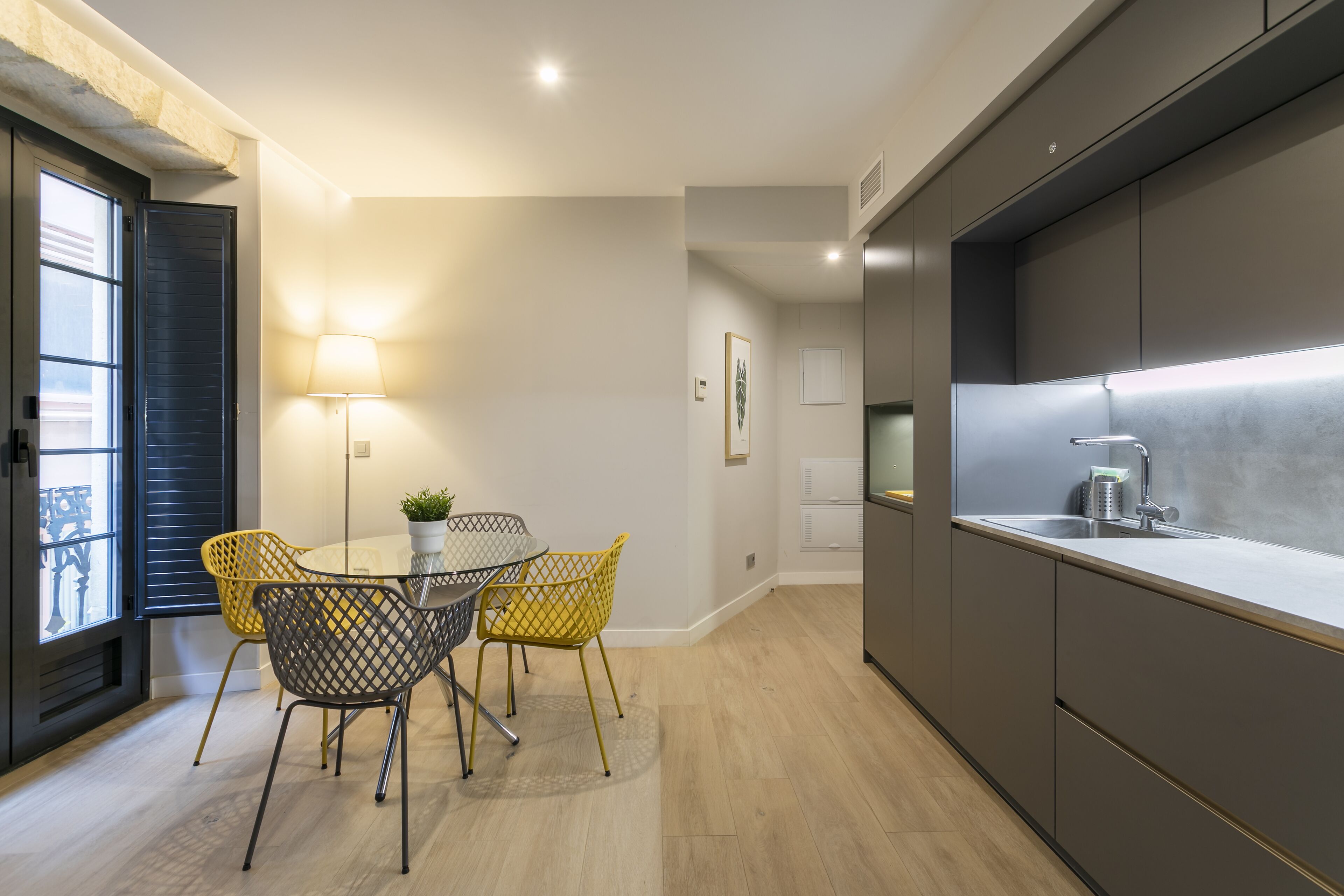 A modern kitchen with a built-in sink, a dining area with a round table and yellow chairs, and a window with blinds.