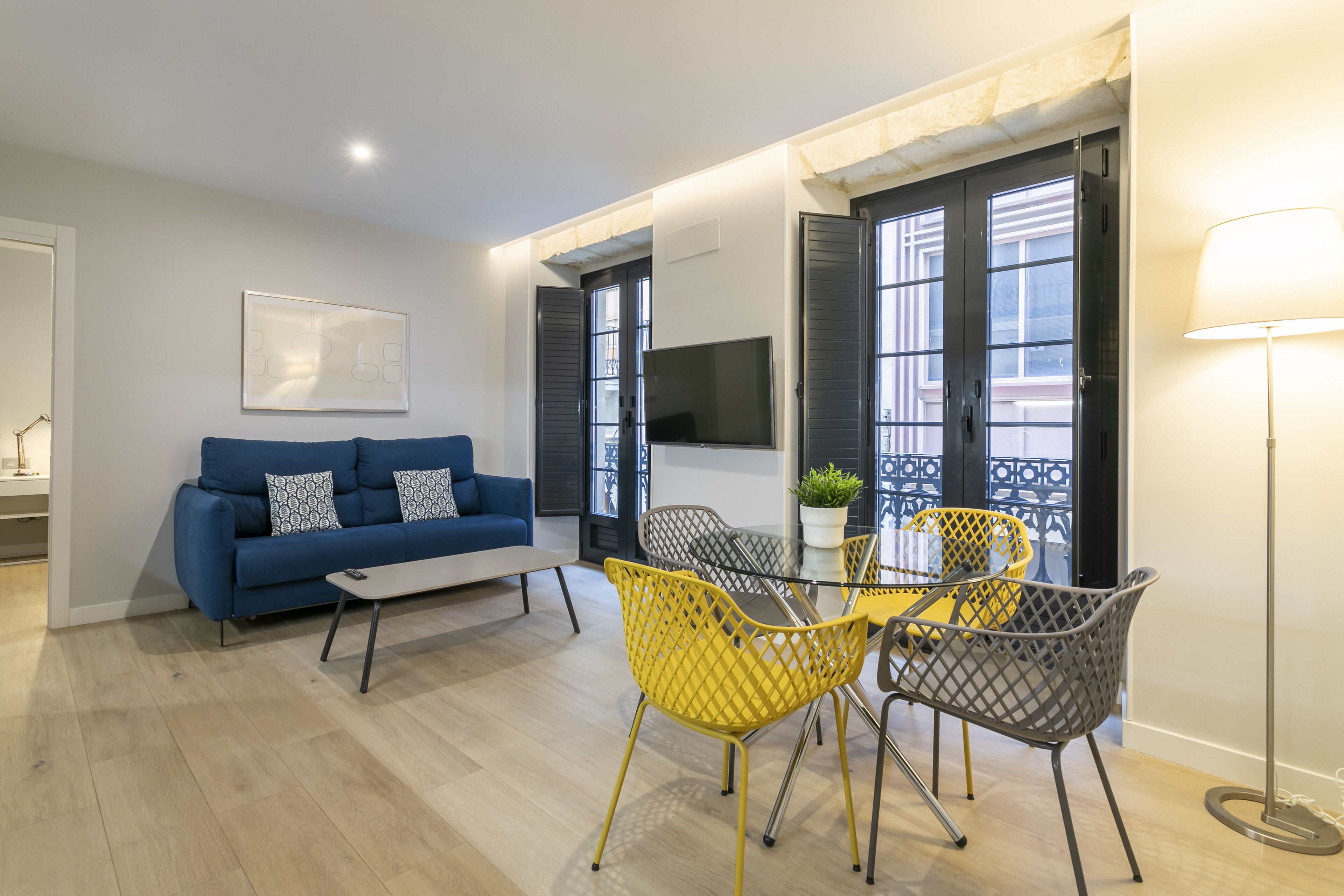Un salon moderne avec un canapé bleu, des fauteuils jaunes, une table basse en verre et un téléviseur fixé au mur.