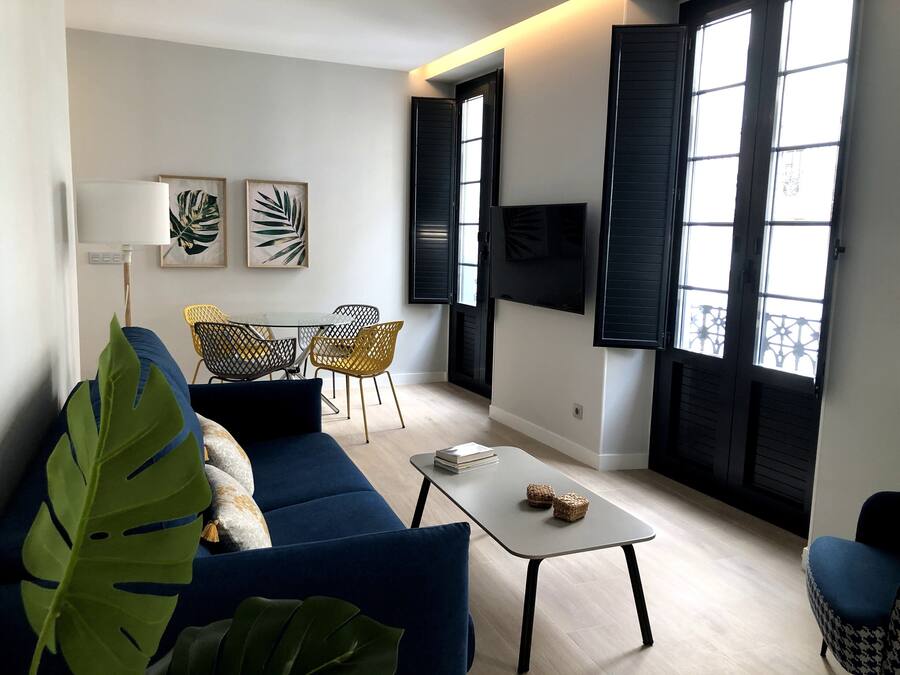 A modern living room with a blue sofa, a coffee table, and a dining area with chairs. There are large windows with shutters, a flat-screen TV, and artwork on the walls.
