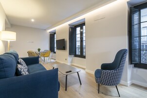 Una sala de estar moderna con un sofá, un sillón y una pequeña mesa. Hay un televisor montado en la pared y una ventana con contraventanas.