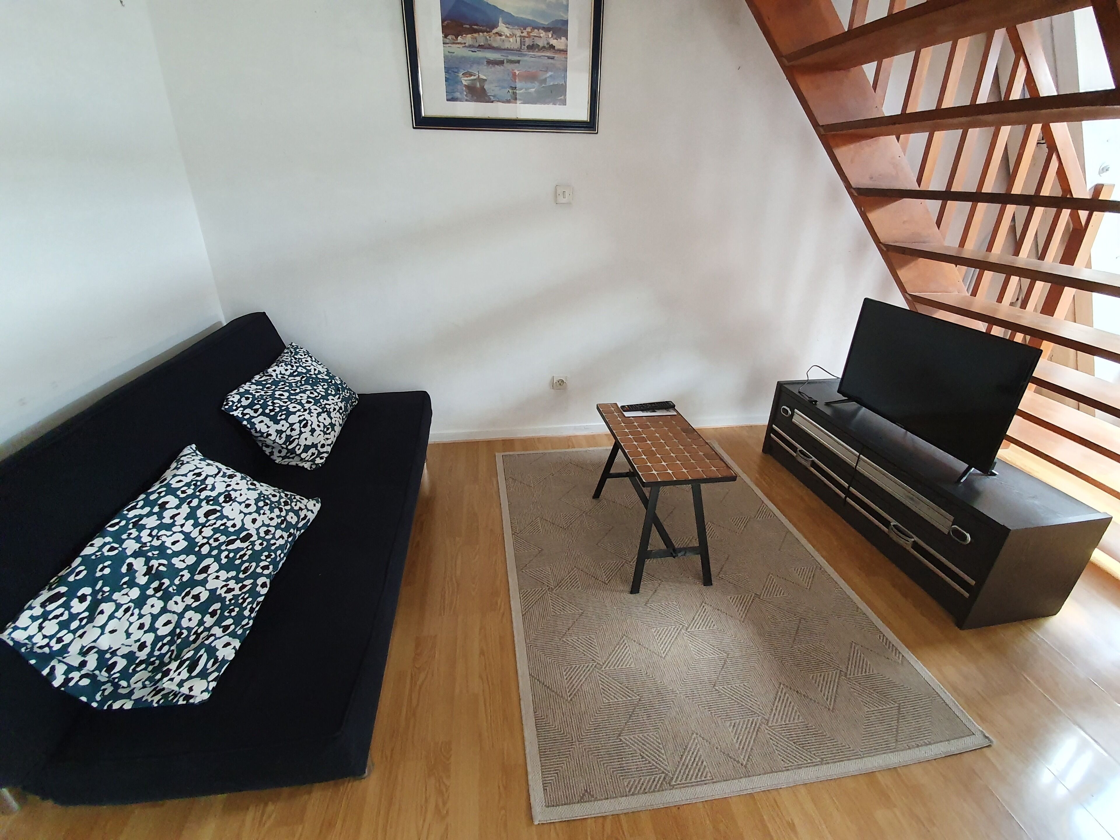 Un salon avec un canapé noir, des coussins à motifs, une petite table en bois, un téléviseur posé sur un meuble et un tapis à motifs.
