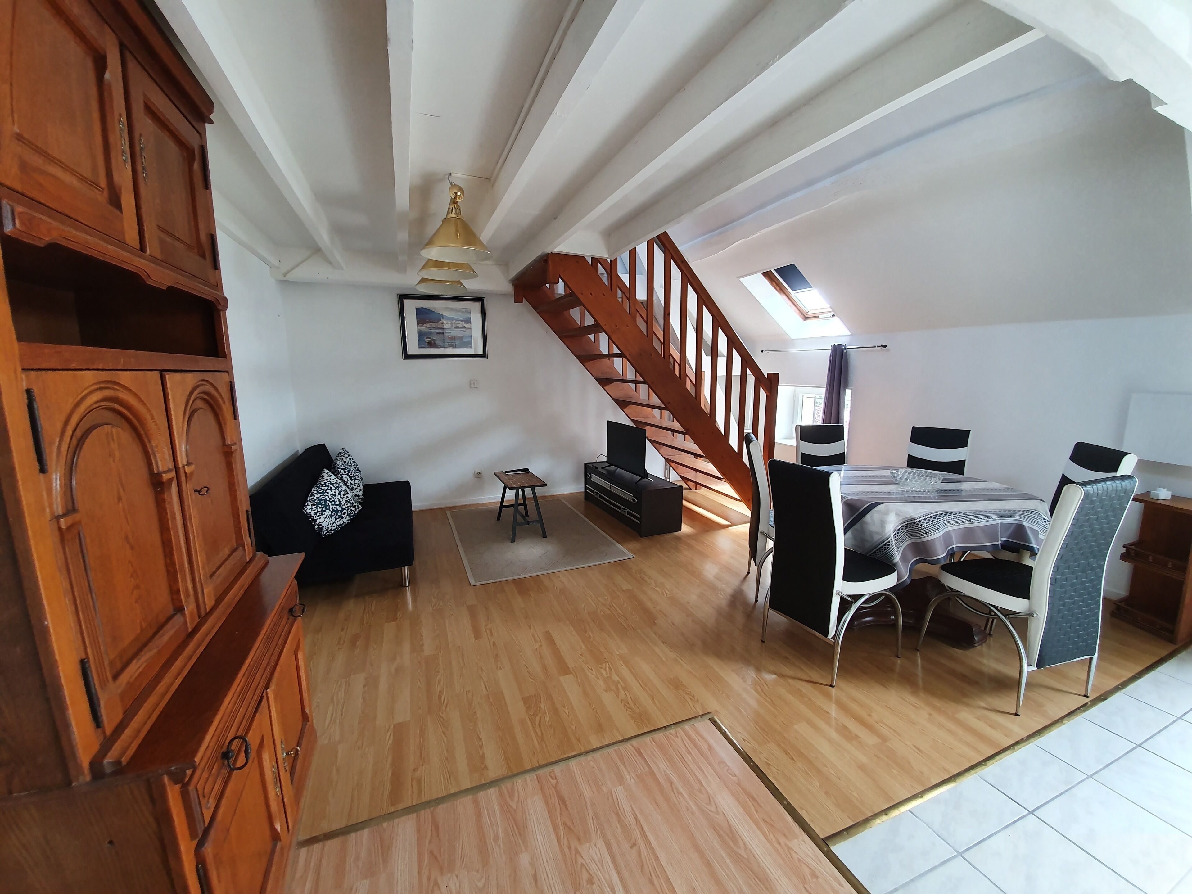 Una habitación amplia con un armario de madera, un sofá negro, una mesa de comedor con sillas y una escalera que conduce a un nivel superior.