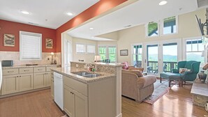 A spacious kitchen with a central island, a dining area with a green upholstered chair, and a view of the outdoors through large windows. - Wintergreens R1231-Mtn Condo/View, Gated Resort, Indoor/Outdoor Pools & Hot Tub (Banner Elk)