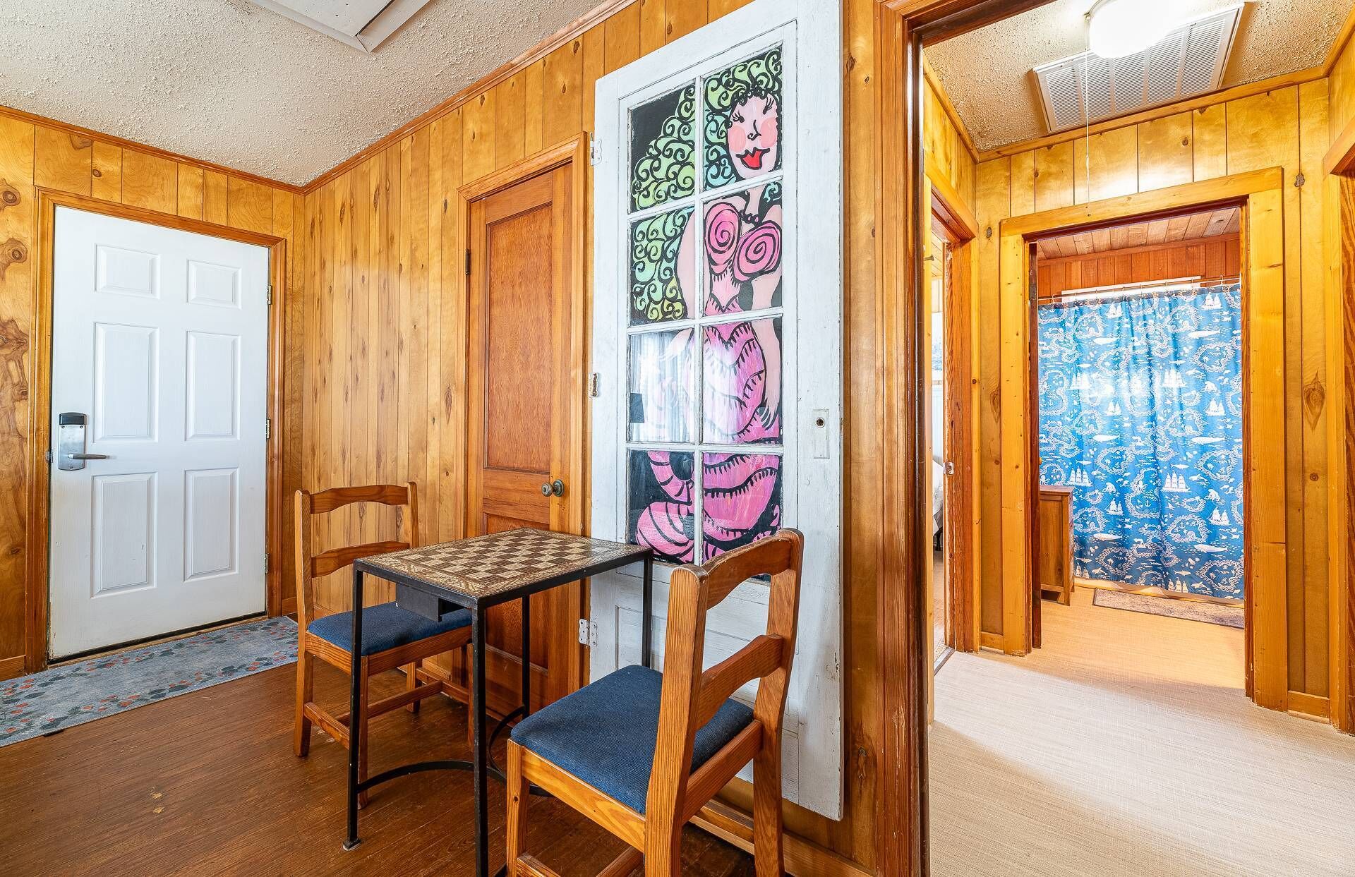 A room with wooden walls, a white door, a chess table, two chairs, and a window with a blue curtain.