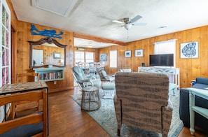 A cozy living room with wooden walls, a ceiling fan, a bookshelf, and a TV. - Beach Baby - Kitty Hawk MP 2.5, Oceanfront, 3 Bedrooms, Best Views on the OBX, Pets OK (Kitty Hawk)