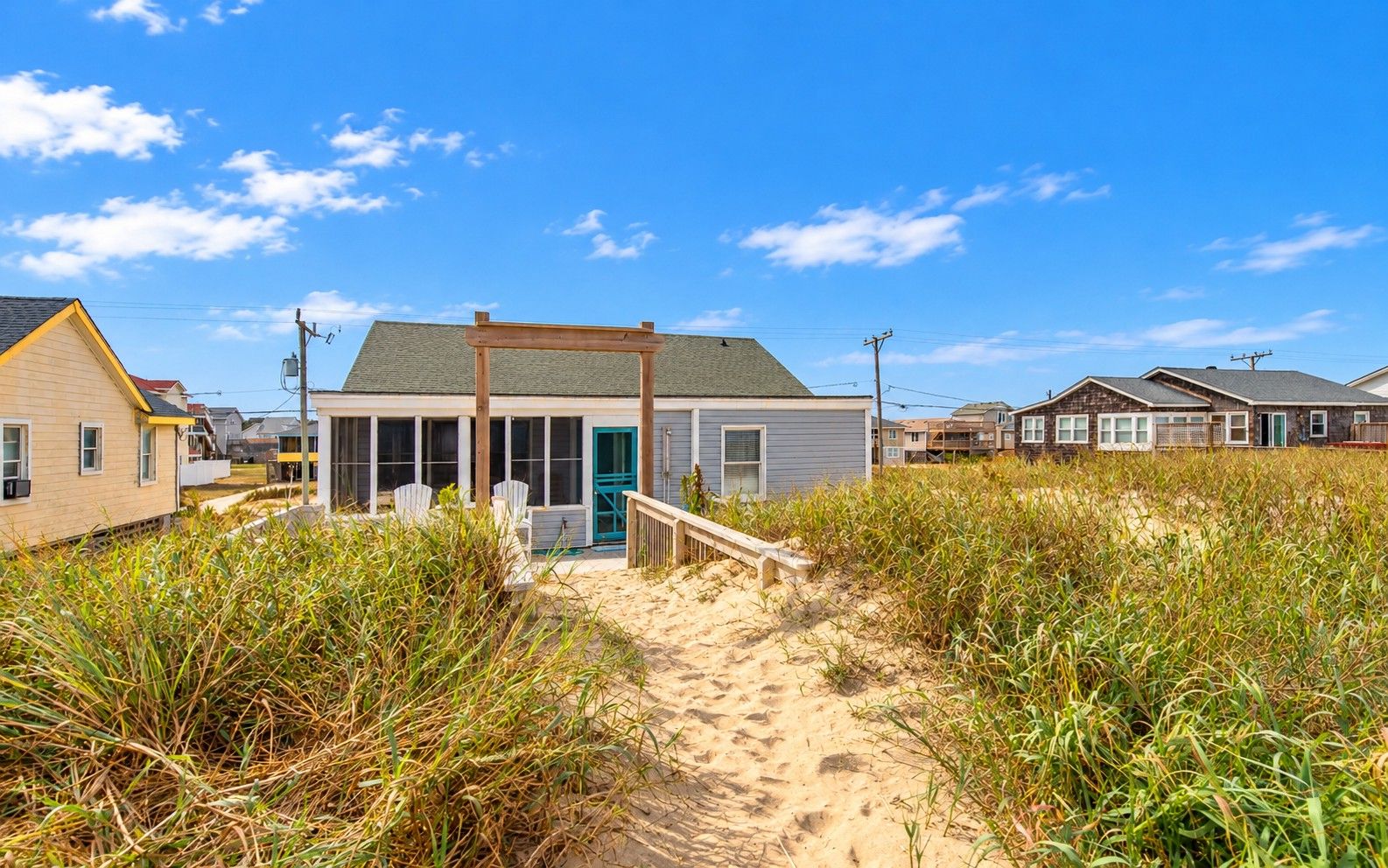 A beachfront property with a covered porch, a sandy path leading to the house, and surrounding vegetation.