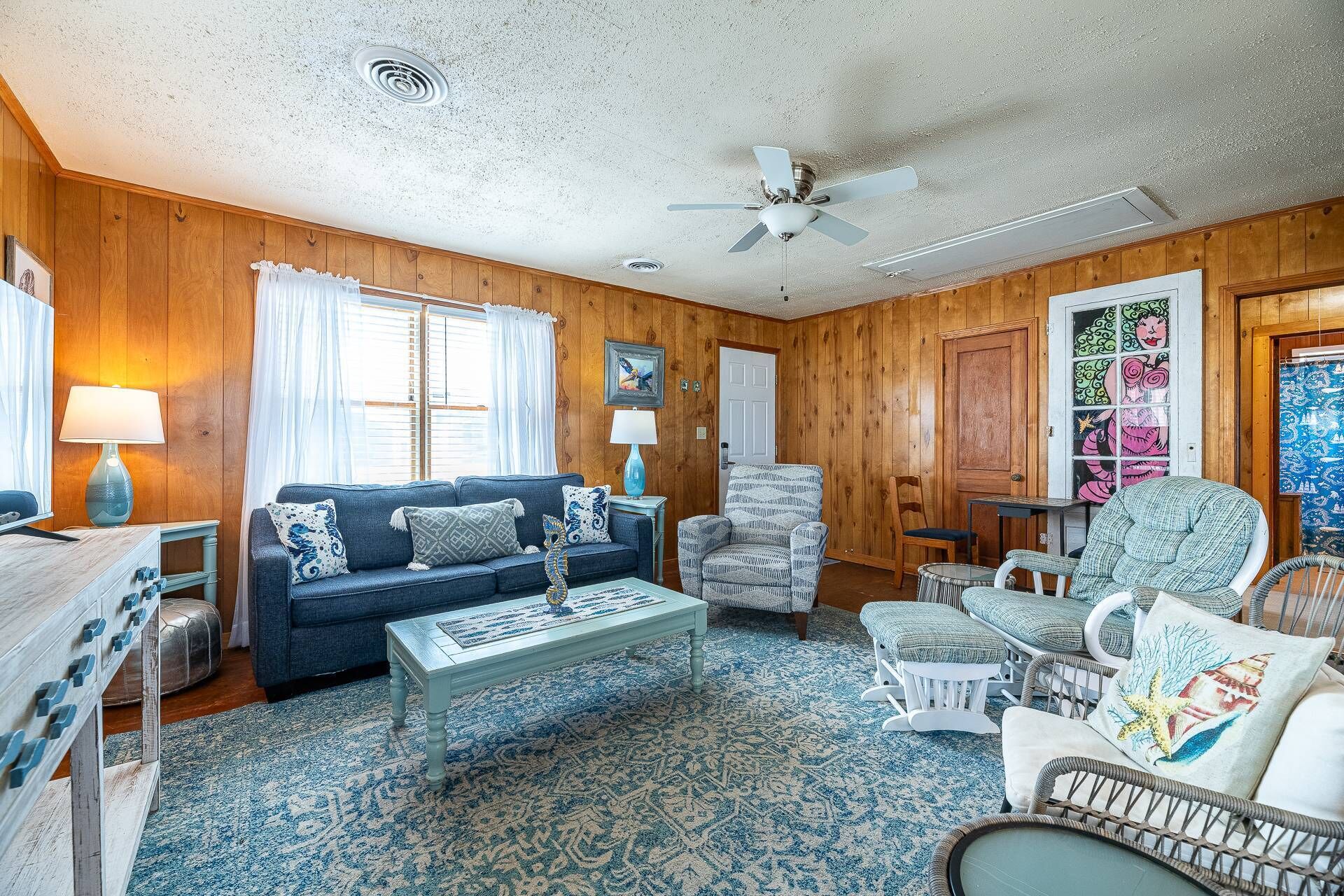 Un salon chaleureux avec un tapis à motifs bleus, des murs en bois, un ventilateur de plafond et un coin repas comprenant une table et des chaises.