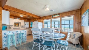 A cozy cabin interior with a kitchen, dining area, and large windows offering a view of the outdoors. - Beach Baby - Kitty Hawk MP 2.5, Oceanfront, 3 Bedrooms, Best Views on the OBX, Pets OK (Kitty Hawk)