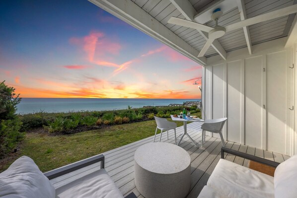 A deck with a round table and chairs, overlooking a grassy area and the ocean at sunset.