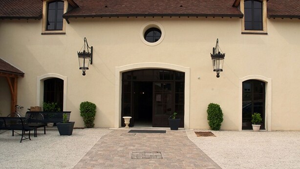A building with a central entrance, two side entrances, and a small circular window above the door. There are two benches and potted plants on the ground. - Le Clos Margot (Blancs-Coteaux)
