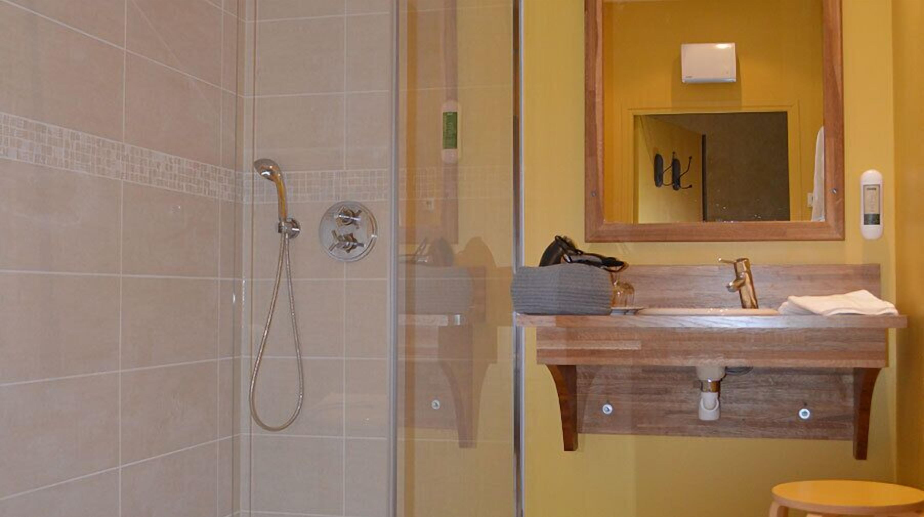 Ein Badezimmer mit einer gläsernen Duschkabine, einem Holzmöbelkorpus mit Spiegel und einem kleinen Hocker.