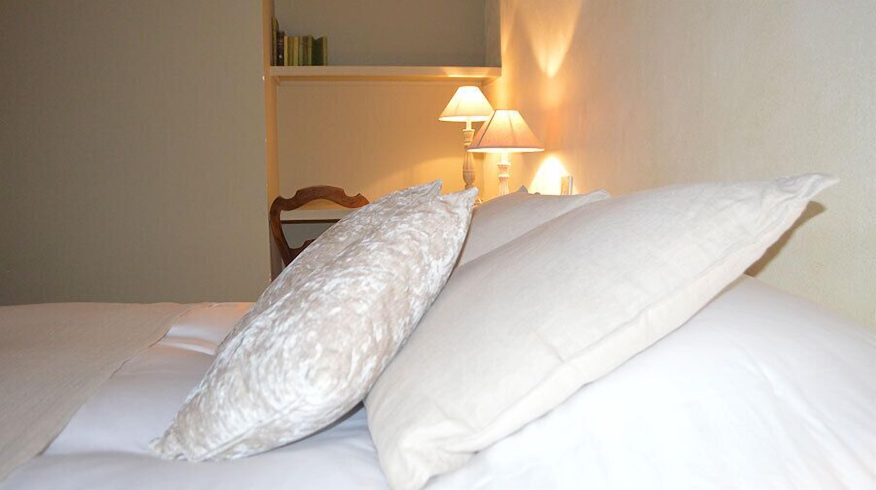 A neatly made bed with white linens and pillows, a wooden headboard, and a bedside lamp.