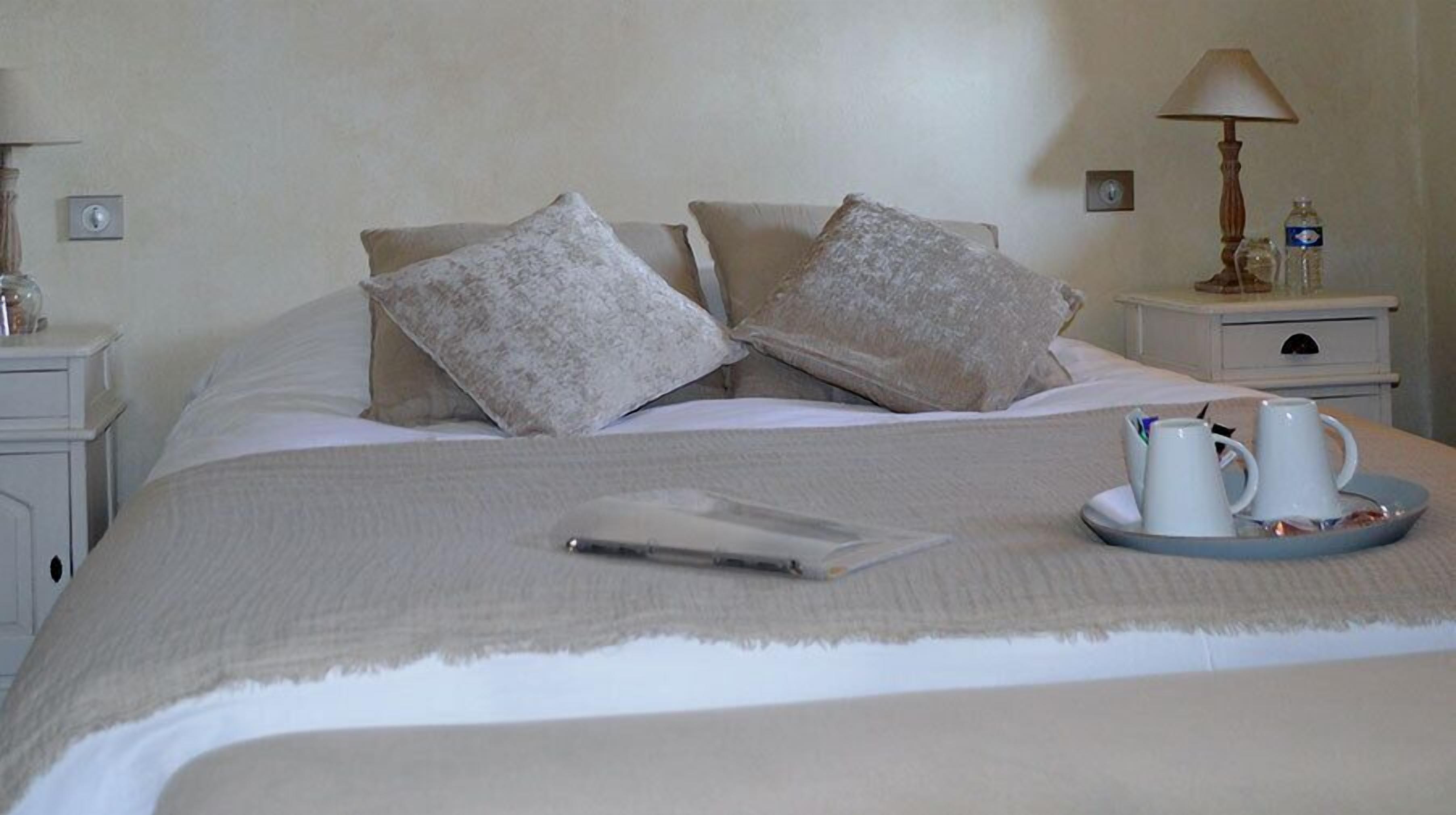 Una cama bien tendida con una bandeja de tazas blancas, una computadora portátil, una lámpara de noche y una botella de agua en la mesita de noche.