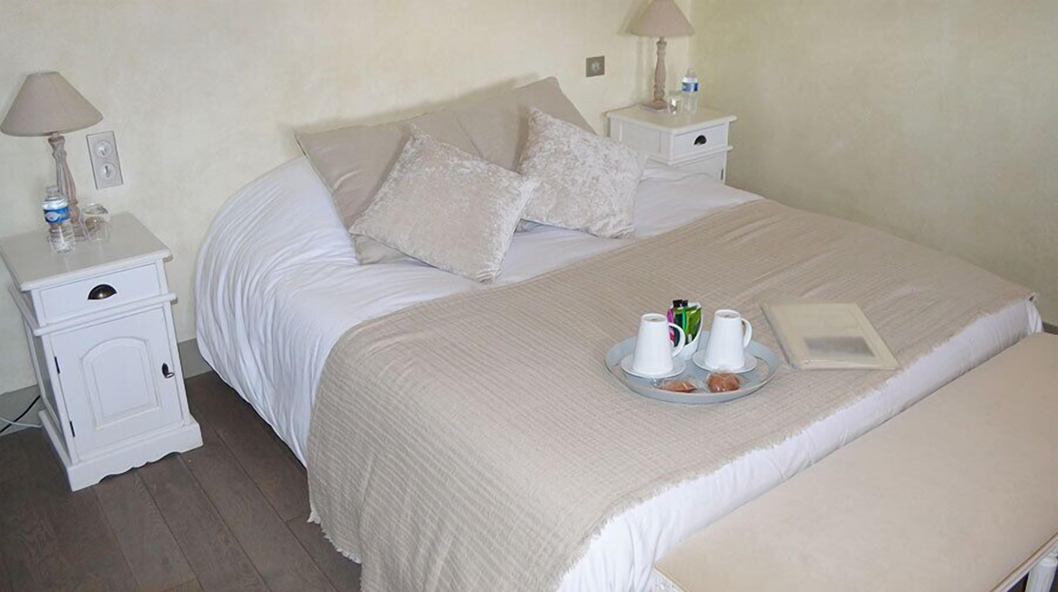 Cama bem arrumada com uma bandeja de itens de café da manhã, incluindo croissants e xícaras de café, em um quarto simples com móveis brancos e um banquinho.