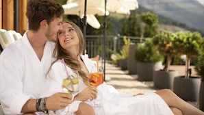 A couple relaxing on a terrace with outdoor seating and umbrellas, overlooking a mountainous landscape.