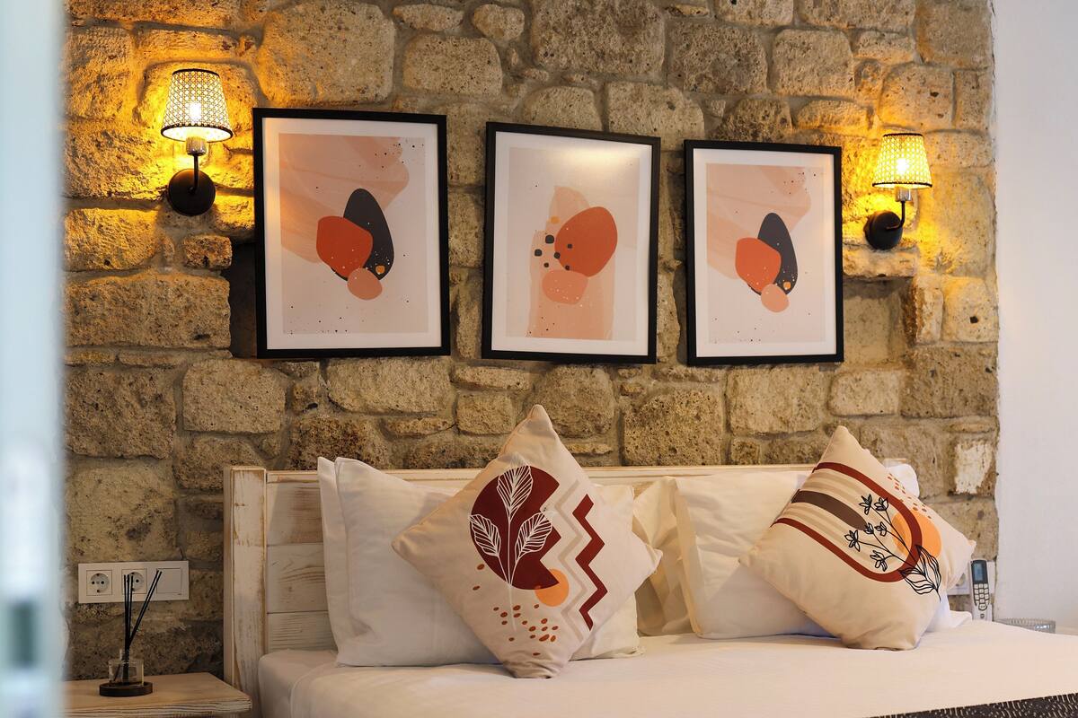 Un lit surmonté de trois œuvres d’art abstraites encadrées, une table de chevet avec une lampe et un mur à la texture pierreuse.
