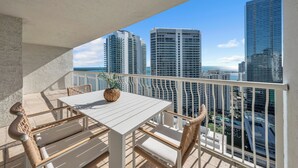 A balcony with a white table and wicker chairs, overlooking a cityscape with tall buildings.