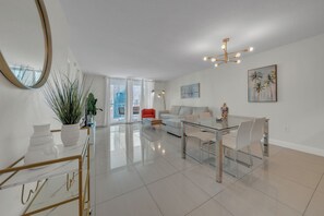 Un salon moderne avec une table à manger en verre, des canapés blancs, un fauteuil rouge, un grand miroir et une vue sur la ville à travers la porte vitrée.